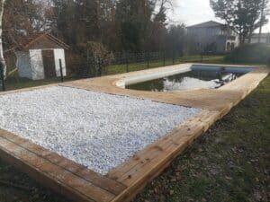 Plage de piscine en bois avec galets blancs, eau sombre reflétant les arbres nus et un cabanon blanc à l'arrière-plan.