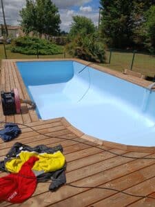 Remplissage d'une piscine neuve avec liner bleu clair posé sur un deck en bois, sous un ciel nuageux. Vêtements "ECOLE" rouges en premier plan.