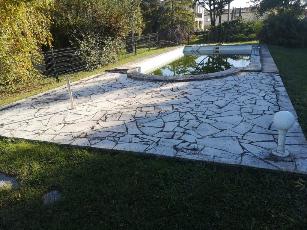 Piscine extérieure partiellement remplie d'eau trouble, entourée d'une terrasse en dalles irrégulières et de gazon, avec des arbustes en arrière-plan.