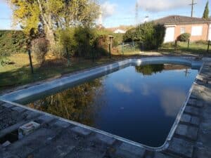 Piscine en cours de rénovation, remplie d'eau sombre reflétant les arbres d'automne et un pylône, avec des travaux visibles sur le pourtour.