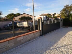 Portail coulissant neuf, mur de parpaings avec chapeaux de pilier rouges, allée gravier blanc. Camion Renault 'Création Aménagement' visible. Ciel bleu.