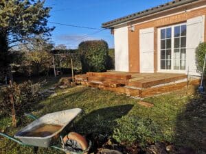 Construction d'une terrasse en bois surélevée devant une maison en briques rouges et crépi blanc ; une brouette est au premier plan.